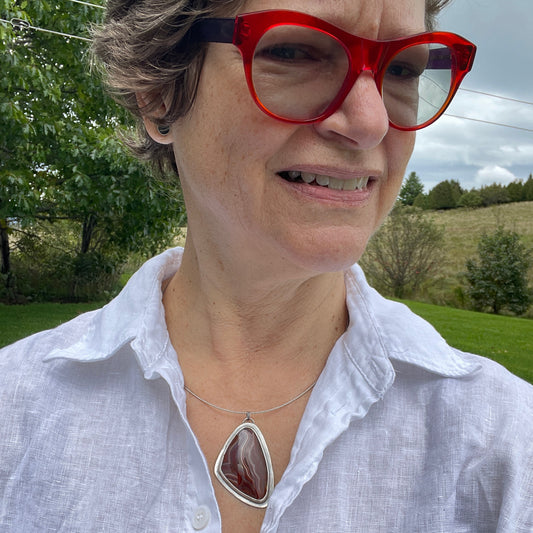 A photo of a white woman with short hair, red glasses and a handmade pendant on a white shirt.